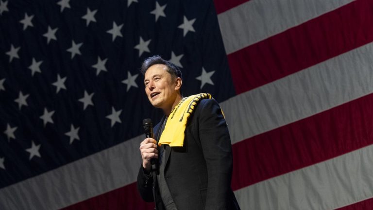 Elon Musk stands in front of a giant American flag