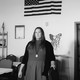 A black-and-white photo of a white woman standing in a room with the American flag behind her