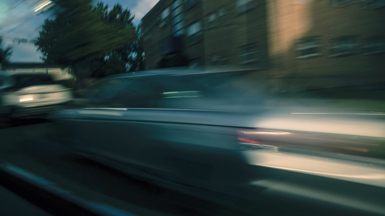 photo of blurred silver car in motion with building and tree in background