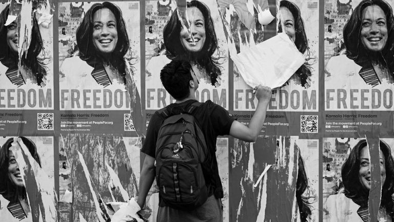 A man tears down a poster among a spread of identical posters depicting Kamala Harris and the word 'freedom.'