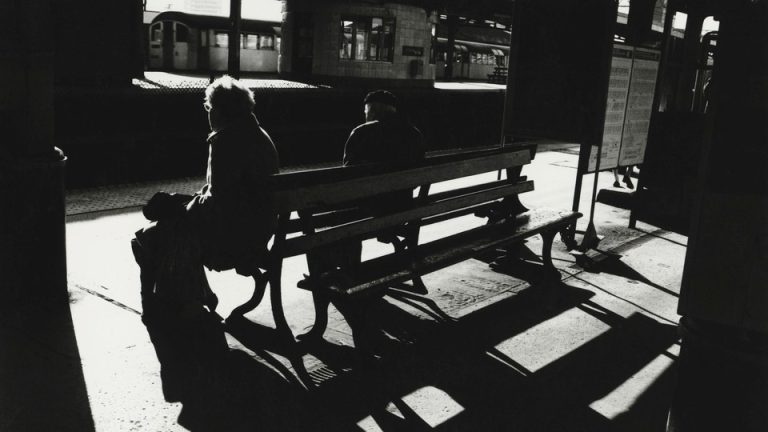 a black and white photo of two people sitting on a bench at a train station, from behind