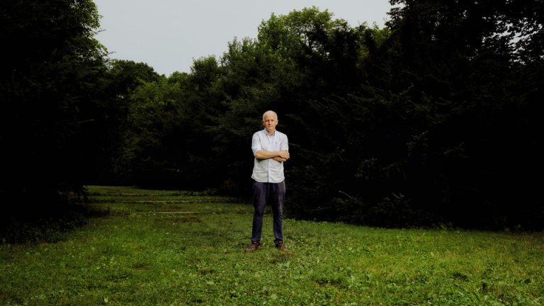 A color photograph of a white man standing with arms crossed on a green lawn surrounded by dark trees