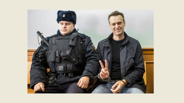 A photo of Alexei Navalny flashing a V sign with his fingers next to a guard