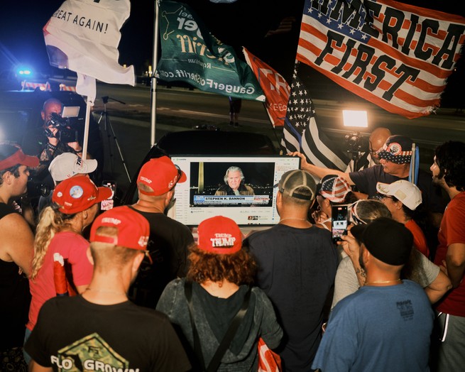 A group of Trump supporters gather around a television screen with Steve Bannon on it.