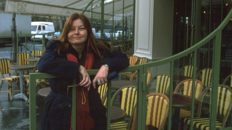 A woman in a blue coat and red scarf with red hair leans on a green railing in an empty outdoor restaurant patio.