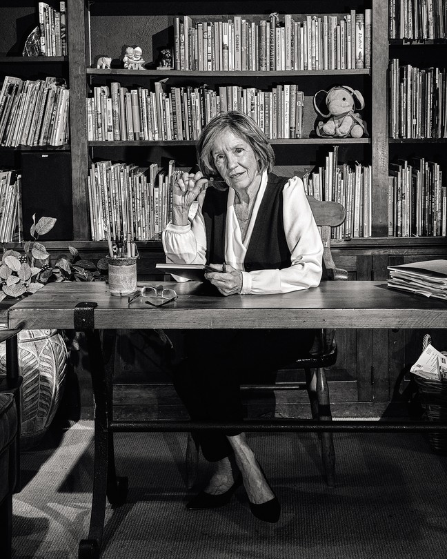 black-and-white photo of woman sitting behind desk next to sofa in home library in front of shelves filled with books