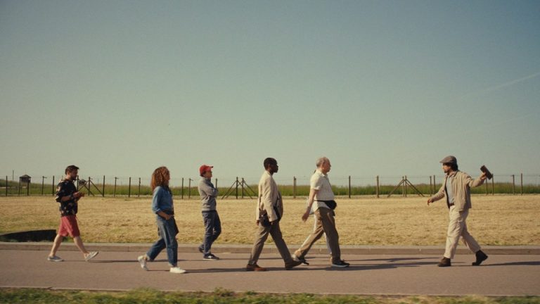 A film still in which a man on the right leads a tour group of five people to his left across a field with barbed wire in the background.