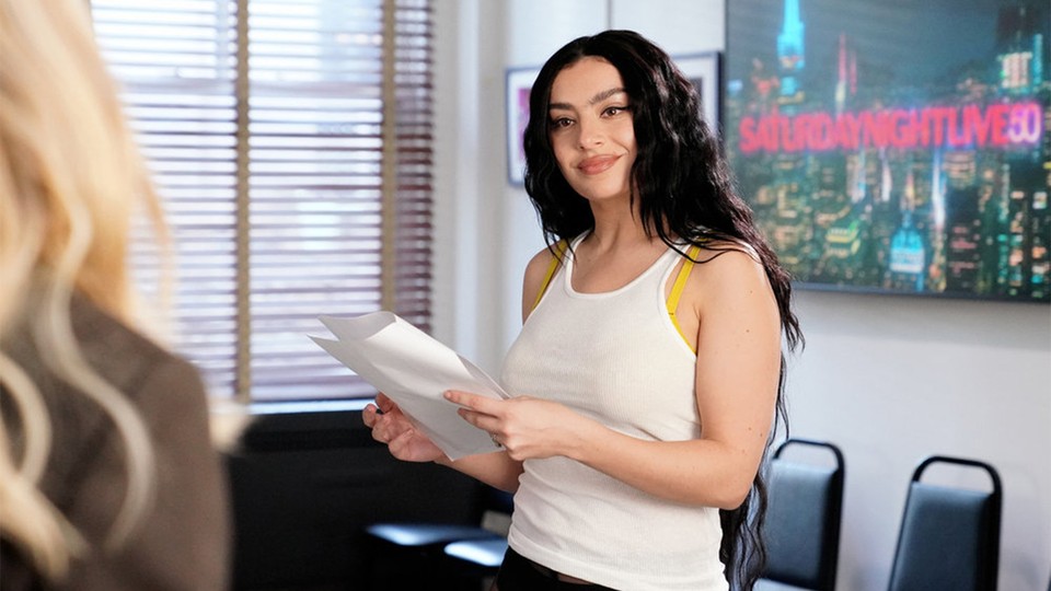 Singer Charli XCX, with long black hair, holds a paper and looks to the left