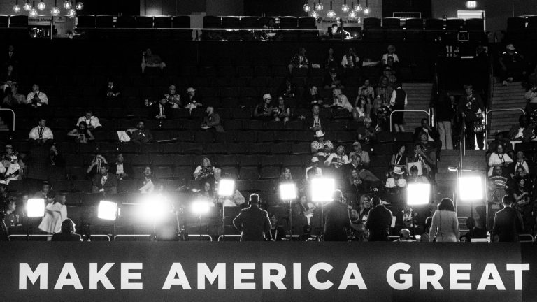 Attendees at the 2024 Republican National Convention sit in the audience, behind a row of bright lights and a banner reading "Make America great"