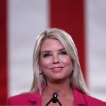 Photo of Pam Bondi smiling at a microphone with a large American flag behind her