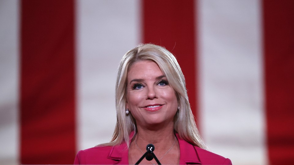 Photo of Pam Bondi smiling at a microphone with a large American flag behind her