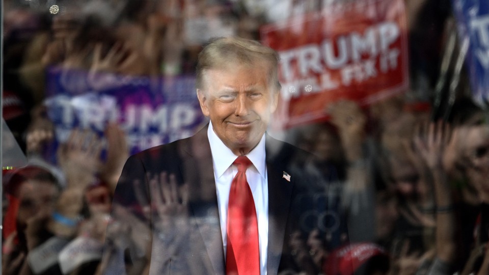 Donald Trump smiles behind a window of bulletproof glass that reflects his cheering supporters and signs