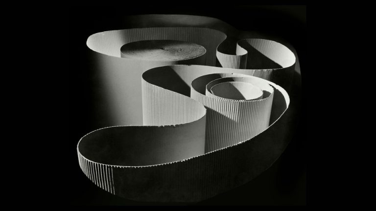 A black-and-white photo of a spotlighted spool of corrugated paper unwinding in artful shapes, surrounded by darkness.