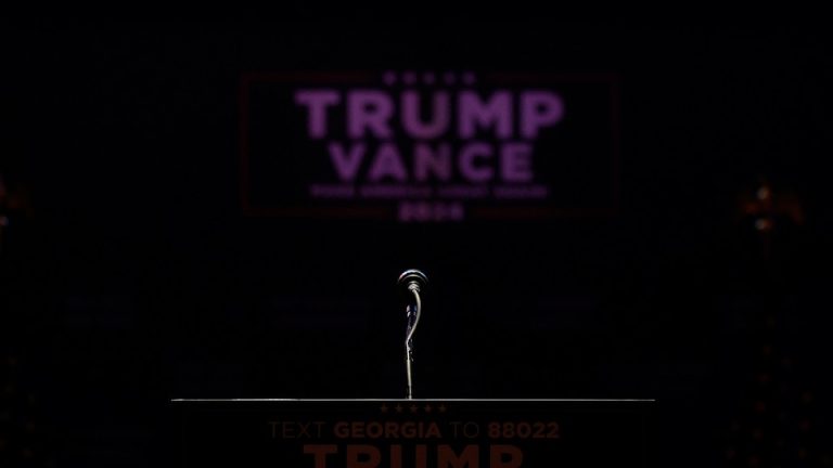 A color photograph of a microphone on a podium, with a black background and a blurred Trump-Vance sign seen in the distance.