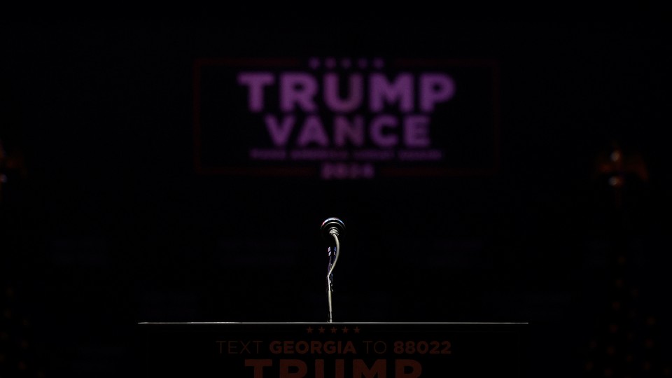 A color photograph of a microphone on a podium, with a black background and a blurred Trump-Vance sign seen in the distance.