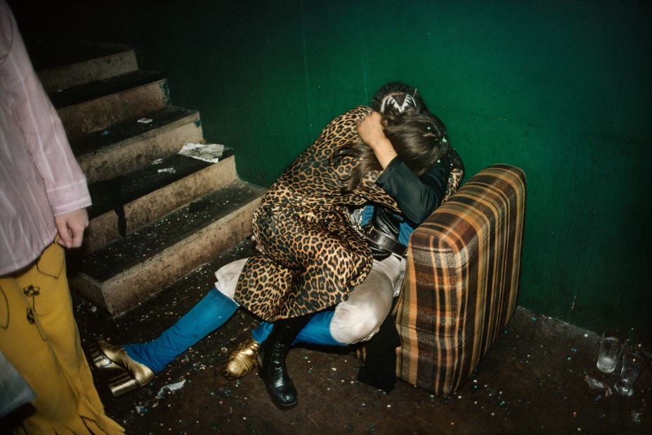 two people hug on a chair on the ground at the base of a staircase. one wears a bright leopard print dress, the other gold heels. another person is in the corner