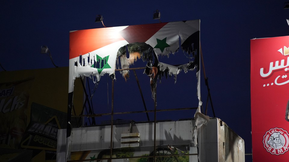 A tattered and torn billboard of the Syrian flag with Bashar al-Assad’s face on it lit up atop a building at night