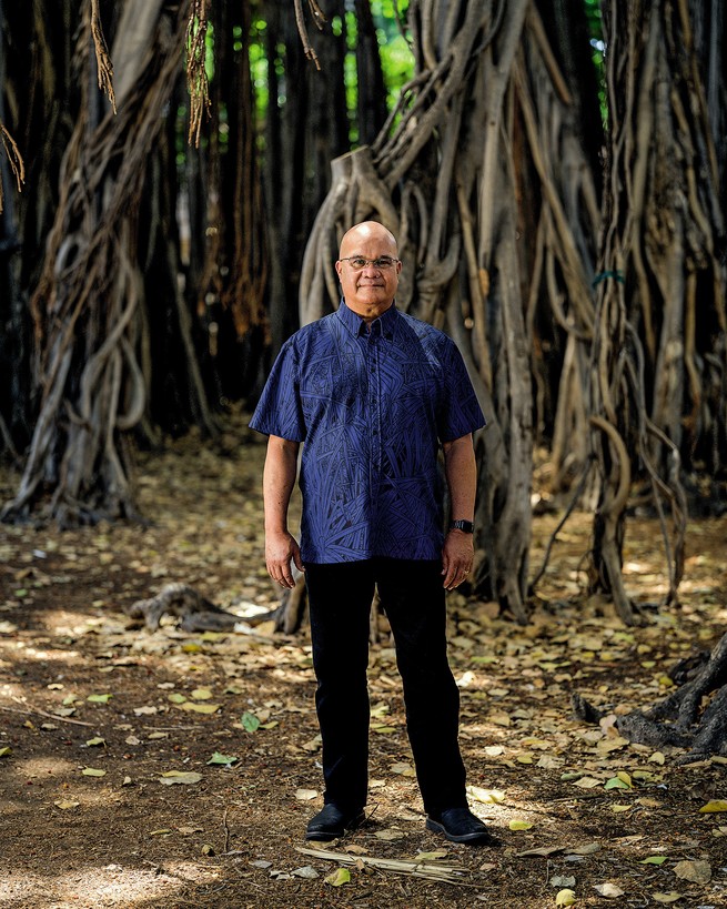 photo of man in short-sleeved blue button-down and black pants with extensive banyan-tree root system in background