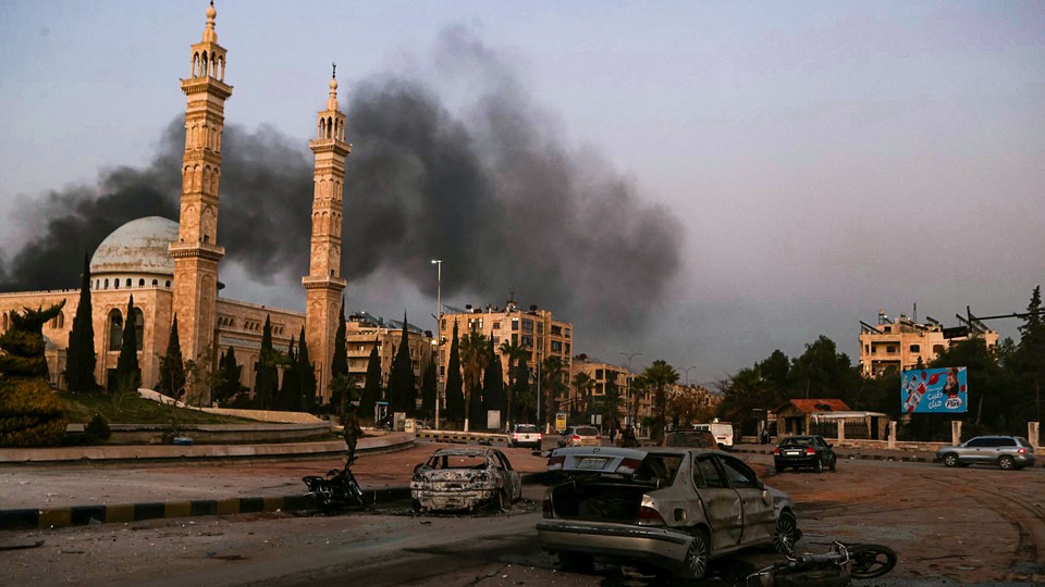 Photo of street view in Aleppo with smoke billowing above buildings and wrecked bombed cars in the foreground.