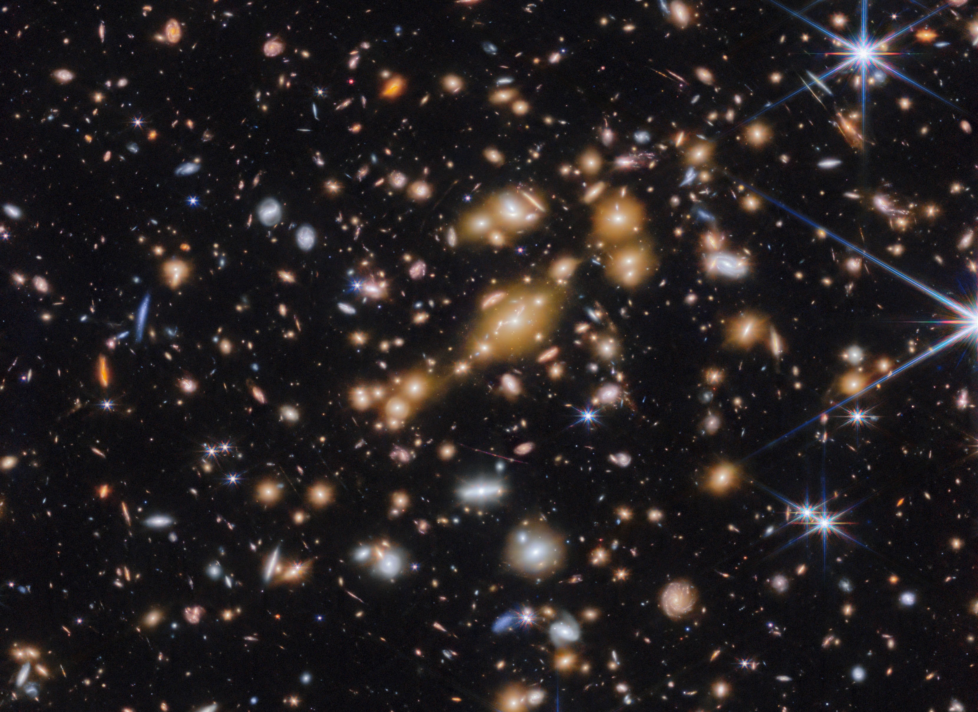 A field of galaxies on the black background of space. In the middle is a collection of dozens of yellowish galaxies that form a foreground galaxy cluster.