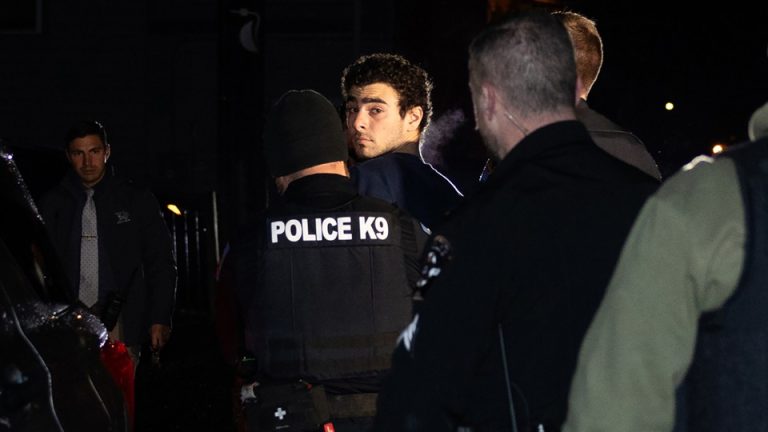 Photo of Luigi Mangione after his arrest looking back at the camera while being escorted away by a cop in a “Police K9” vest