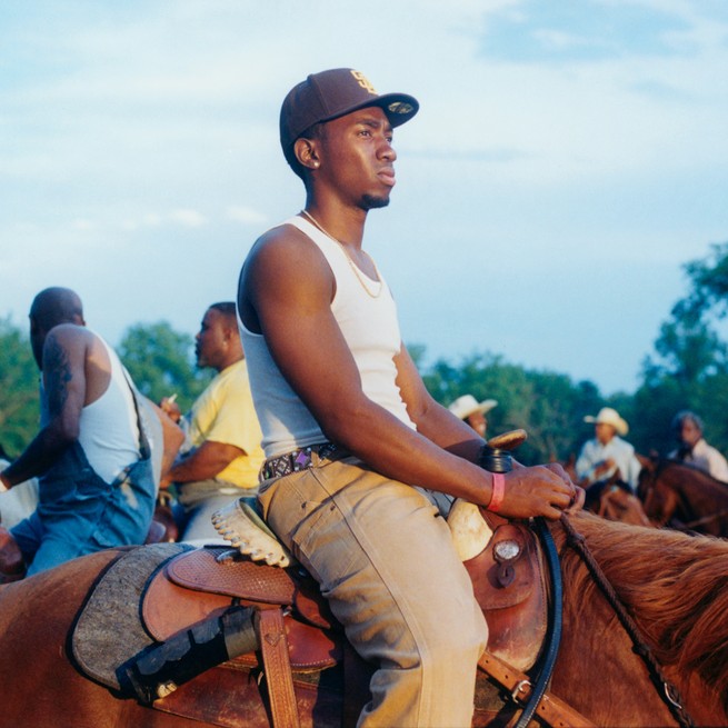 photo of young Black man in baseball cap, white tank, khaki-color pants, mounted on chestnut horse under Western saddle with other riders in background