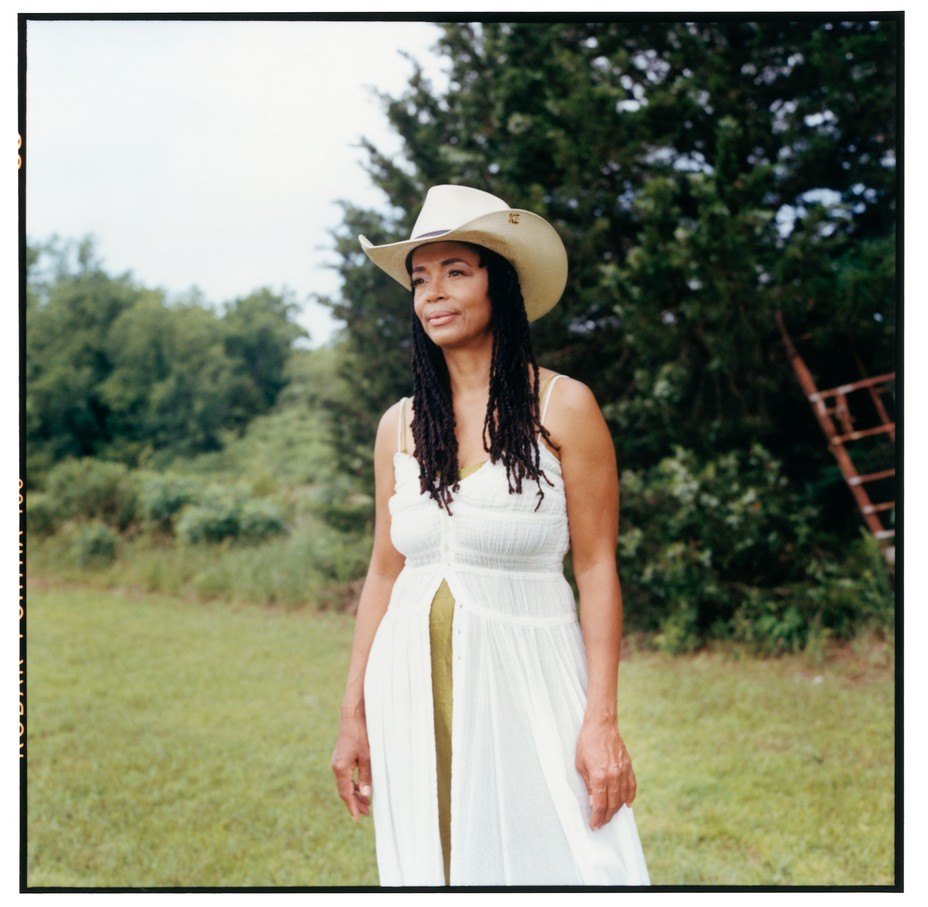 photo of Black woman with long hair wearing gauzy white sundress and straw summer cowboy hat standing in field with fence and trees behind