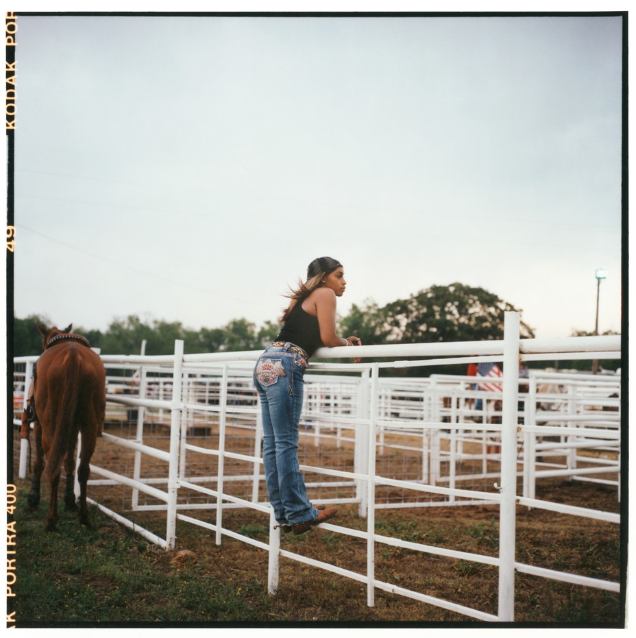 woman with long hair in black tank top, jeans with embroidered back pocket, and spurs stands on white arena fence rail watching, with horse in background hitched to rail