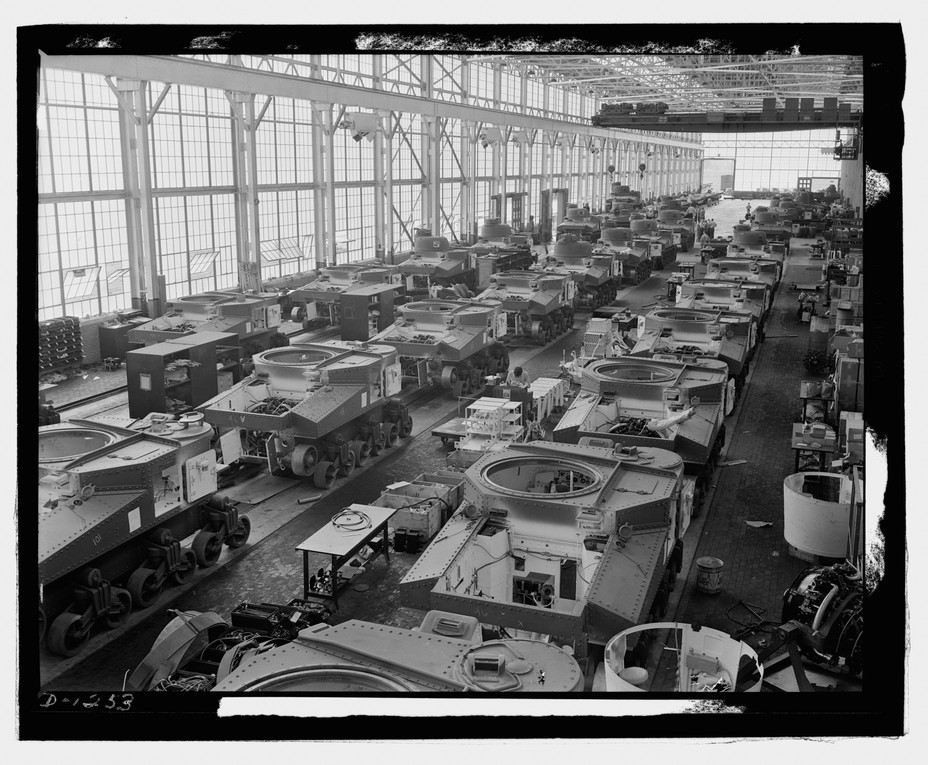 A black and white photo shows an assembly line of tanks lined up in a warehouse.