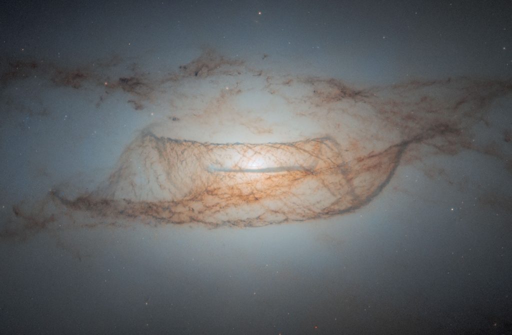 A telescopic view of a hazy galaxy lined with lanes of dust that form a rough wavy pattern