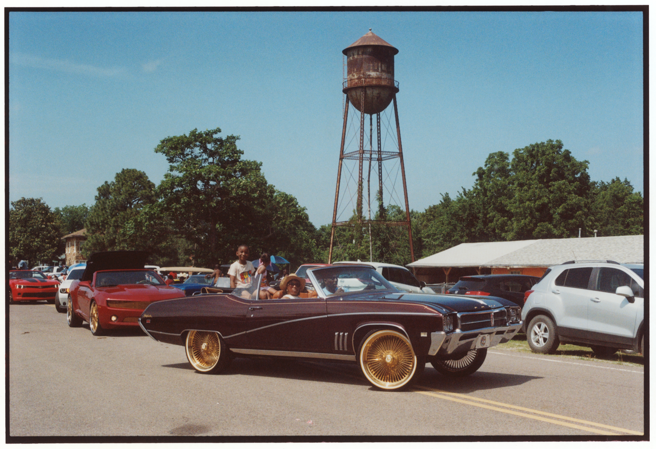 photo of parade of cars driving into rodeo with rusting water tower in background