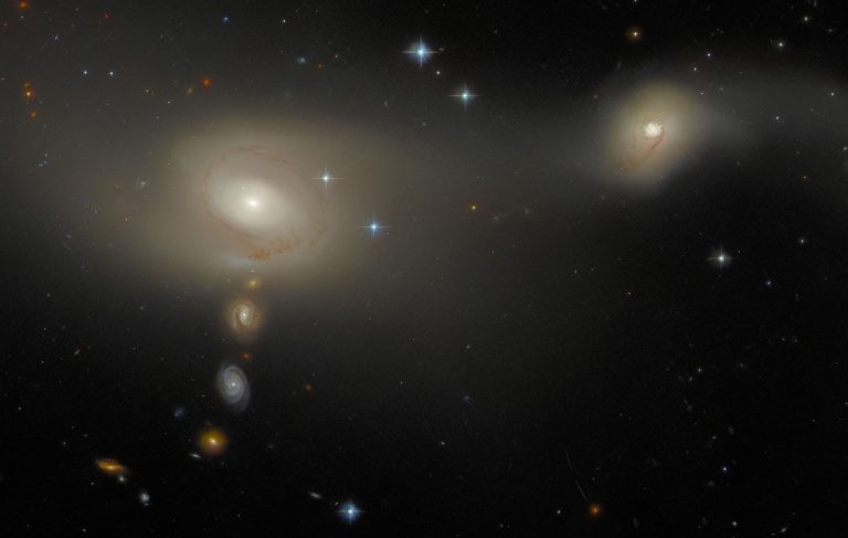 A pair of interacting galaxies, one smaller than the other. They appear as a broad, soft glow in which individual stars can’t be seen. A number of bright stars and smaller, background galaxies can also be seen — three such galaxies lie in a vertical line below the right-hand galaxy of the pair.