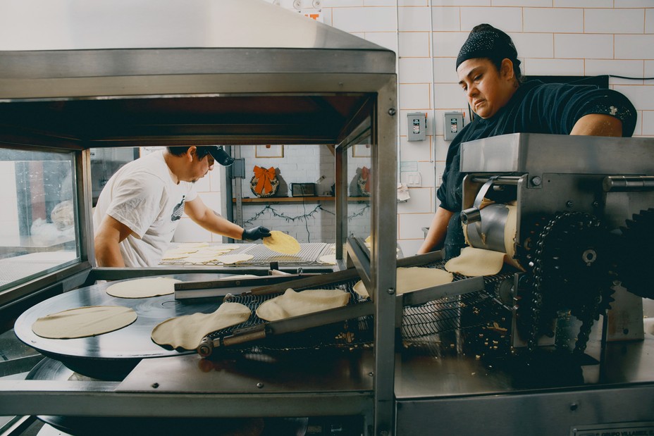 People roll out tortillas on a conveyor belt in a kitchen