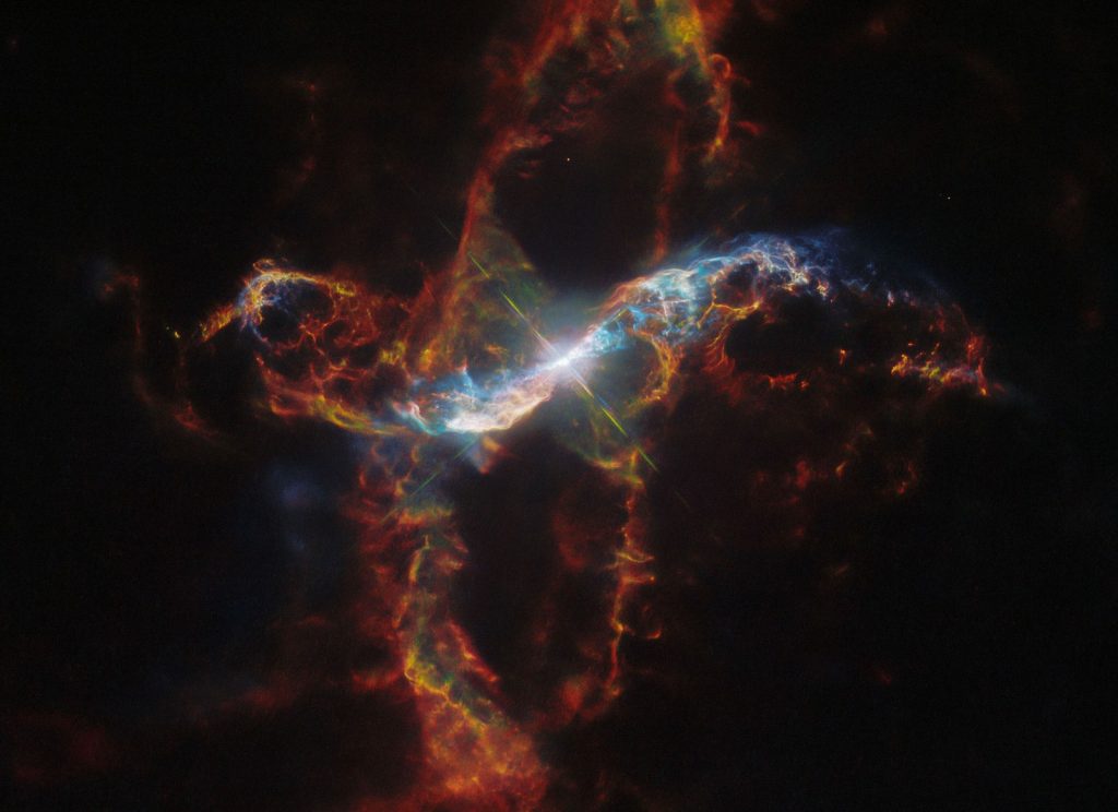 A bright binary star surrounded by a nebula. The star, in the center, is a large white spot surrounded by a circular glow. The nebula extends far above, below, left and right of the star in long, arcing shapes made of thin, multicolored filaments.