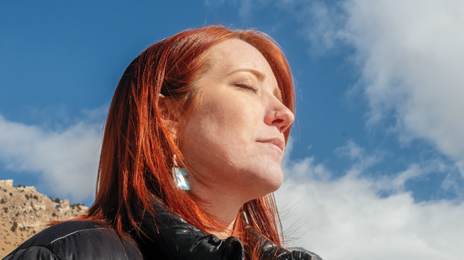 A photograph of red-haired woman with eyes closed wearing black jacket in sunlight with mountain and blue sky in background