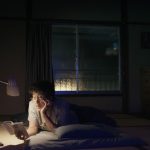 A photograph of a person laying on the floor of a cozy bedroom at night with a lamp illuminating the book the person is reading