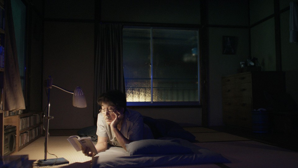 A photograph of a person laying on the floor of a cozy bedroom at night with a lamp illuminating the book the person is reading