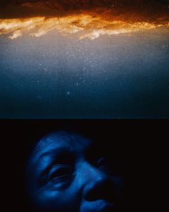 the top half of the photo shows an upside down landscape, with clouds above mountains and a dark starry sky above that. the sky bleeds into an image of a person's face in the dark