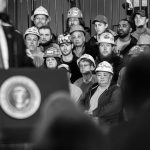 Black and white photo of Donald Trump speaking at a podium with workers wearing hard hats seated behind him.