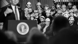 Black and white photo of Donald Trump speaking at a podium with workers wearing hard hats seated behind him.