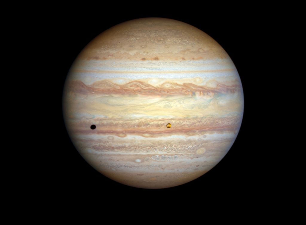 A view of the planet Jupiter, showing many broad and multi-colored bands of swirling storms in its atmosphere, and one small moon that casts a shadow