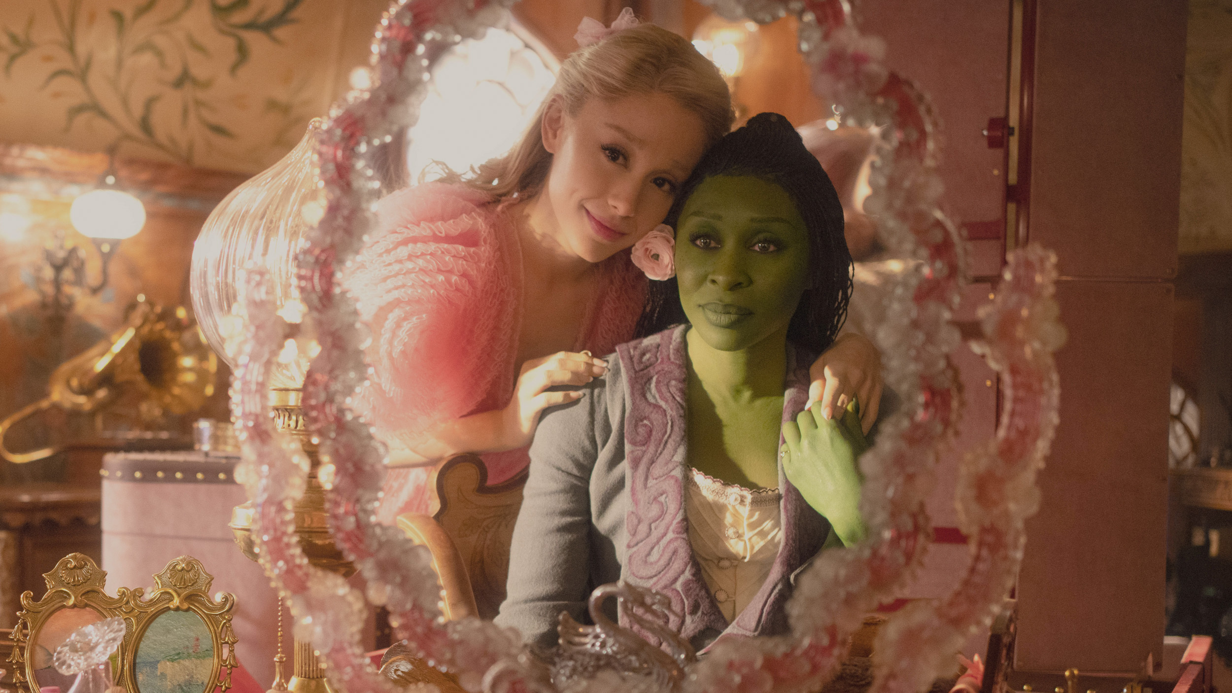 Glinda and Elphaba smile at each other in front of a pink mirror