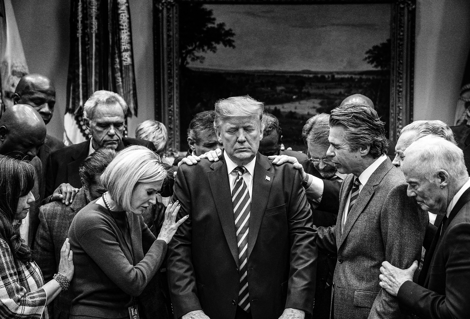 black-and-white photo of Trump standing with eyes closed, surrounded by men and women in suits laying hands on him and praying, with framed oil painting and flags in background