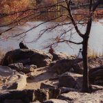 photo of people sitting on rocks next to water with fall foliage