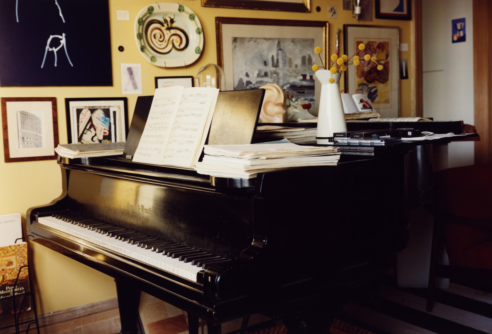 photo of a baby-grand piano with sheet music and art hung on yellow wall behind