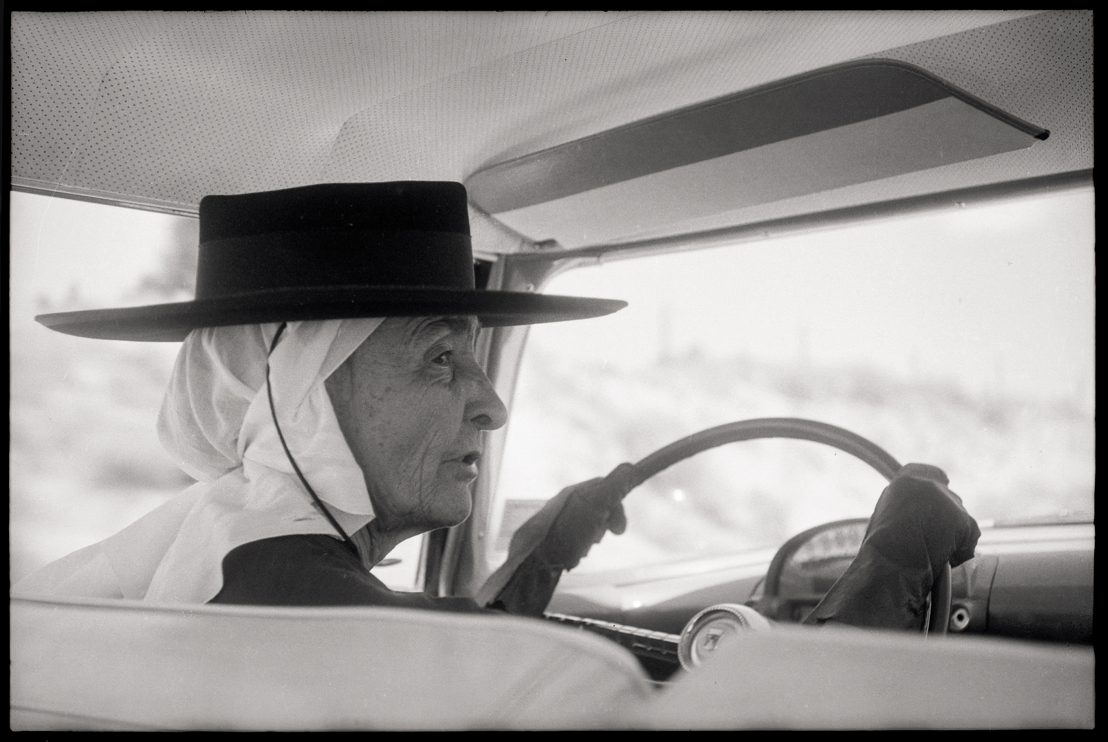 black-and-white photo taken from back seat of O'Keeffe driving car while wearing gloves, scarf over hair, and broad-brimmed black hat