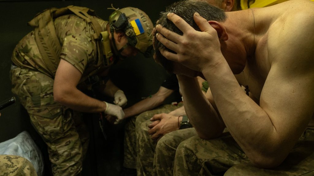 A Ukranian soldier holds his head in his hands while another one behind him treats another soldier.