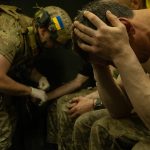 A Ukranian soldier holds his head in his hands while another one behind him treats another soldier.